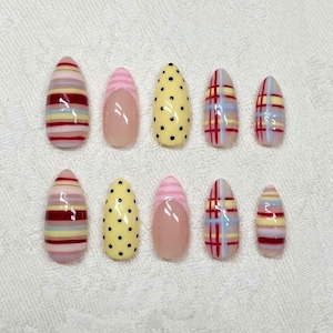 May include: A collection of ten false nails with diverse patterns. Some nails display horizontal stripes in red, yellow, and pink. Others have a yellow base with black polka dots, or a pink base with a pink stripe. Two nails have a plaid design.