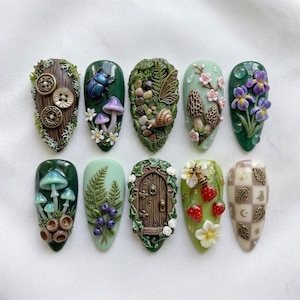 May include: A collection of ten intricately designed nail art pieces. Each nail features a unique, nature-inspired design, including mushrooms, beetles, flowers, and a miniature fairy door. The color palette includes greens, browns, and pastels, with detailed textures.