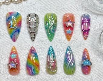 Op de afbeelding: Een verzameling van tien lange, puntige kunstnagels. Ontwerpen omvatten regenboogwervelingen, zilveren versieringen en ster- en hartaccenten. Kleuren variëren van levendige regenboogtinten tot iriserende groen- en blauwtinten.