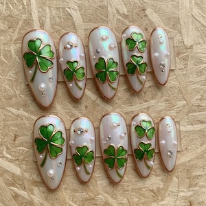 Könnte beinhalten: Zehn künstliche, ovale Fingernägel mit Perlmutt-Finish. Jeder Nagel zeigt ein grünes Kleeblatt-Design mit goldenen Verzierungen, verziert mit Perlen und Strasssteinen. Perfekt für St. Patrick's Day.
