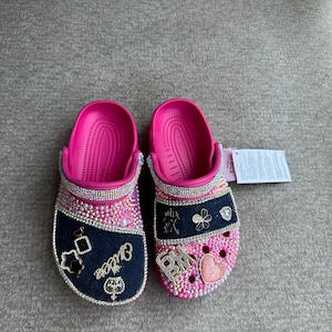 Könnte beinhalten: Ein Paar pinkfarbene Crocs, verziert mit Strasssteinen und Charms. Ein Schuh hat einen dunkelblauen Denim-Patch mit dem Wort "Cuties" und verschiedenen Charms. Der andere Schuh hat einen rosa Herz-Charm und die Initialen "BM".