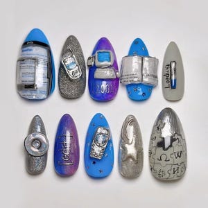 May include: A collection of ten almond-shaped artificial nails, each adorned with unique 3D designs. The nails showcase miniature models of a computer, a mobile phone, and a book, alongside the word "Loading" and other text. The colour scheme includes blue, silver, and purple.