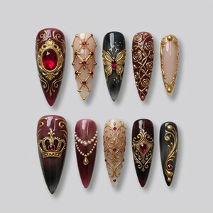 May include: Ten long, almond-shaped artificial nails with elaborate designs. Each nail showcases gold detailing, red gems, and ornate patterns. The colour scheme includes burgundy, black, gold, and nude tones, creating a luxurious aesthetic. The nails are approximately 2.5 cm long.