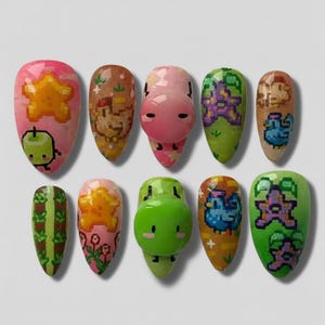 Puede incluir: Diez u&ntilde;as en forma de almendra pintadas con dise&ntilde;os &uacute;nicos. Las u&ntilde;as presentan estrellas pixeladas, manzanas y personajes de dibujos animados en tonos rosa, verde, marr&oacute;n y azul. Los dise&ntilde;os son detallados y caprichosos, mostrando una variedad de estilos.