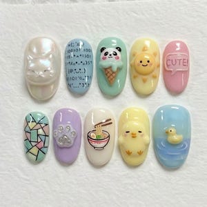 Può includere: Una collezione di dieci nail art. I disegni includono un gatto, codice binario, un panda con gelato, un sole sorridente, la parola "CUTE!", un motivo a vetrata, un'impronta di zampa, una ciotola di ramen, un pulcino e un'anatra nell'acqua.