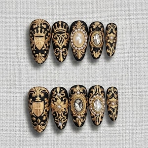 May include: A collection of black press-on nails with elaborate gold detailing. Each nail showcases a unique design, such as shields, crowns, and floral patterns, some with pearl and crystal embellishments. The oval-shaped nails are displayed on a neutral background.