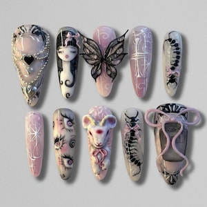 May include: A collection of ten long, almond-shaped artificial nails. Each nail features unique designs, including a black butterfly, a girl's face, a heart, and other intricate patterns in pink, black, and white. The nails are arranged on a light gray surface.