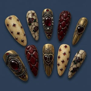 May include: A collection of ornate, almond-shaped nail art designs. The nails feature a variety of embellishments, including red gems, quilted textures, and floral and heart motifs. The color palette includes cream, gold, and burgundy.