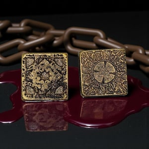 May include: Two gold-coloured puzzle boxes with detailed patterns, one with a sunburst design and the other with an ornate floral pattern. A brown chain is in the background, and a pool of red liquid is in the foreground.