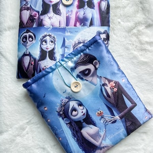 Puede incluir: Dos bolsas de tela azules con ilustraciones de personajes de la película animada "La novia cadáver". Las bolsas tienen un cierre de botón con un cordón turquesa. Las ilustraciones incluyen a la novia, el novio y otros personajes en una escena de boda.