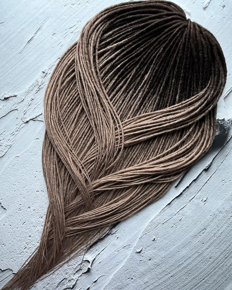 May include: A set of long, brown ombre synthetic dreadlocks. The dreadlocks are arranged in a loose braid and have a light brown color at the roots that gradually transitions to a darker brown color at the ends.