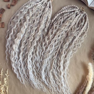 May include: Two sets of wavy, light blonde synthetic dreadlocks, arranged to resemble a heart shape. The dreadlocks are a mix of light blonde and beige tones. The image is taken on a brown surface, with decorative elements like wood chips and dried plants.