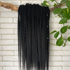 Peut inclure: Des dreadlocks synthétiques noirs suspendus à une branche contre un mur de briques blanches. Les dreadlocks sont longues et droites.