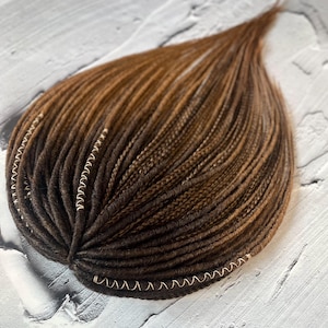 Puede incluir: Un paquete de rastas sintéticas marrones y marrones claras con cuentas doradas en los extremos.