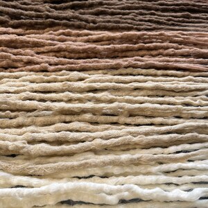 May include: A collection of synthetic dreadlocks in various shades of brown, beige, and white. The dreadlocks are arranged in rows, creating a gradient effect.
