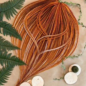 May include: A bundle of long, synthetic hair extensions in shades of orange, brown, and white. The hair is arranged in a loose, flowing style.
