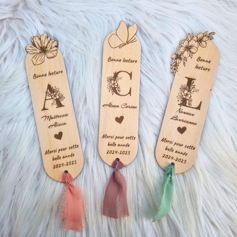 May include: Three wooden bookmarks with floral and butterfly designs. Each bookmark has a different initial, name, and the text "Bonne lecture" and "Merci pour cette belle ann&eacute;e 2024-2025". Each bookmark has a ribbon attached.