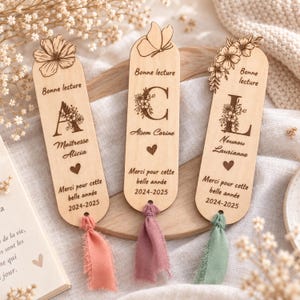 Peut inclure: Trois marque-pages en bois clair aux extrémités arrondies, chacun gravé au laser avec une lettre, des motifs floraux et le texte "Bonne lecture", "Merci pour cette belle année" et "2024-2025". Chaque marque-page possède un pompon en tissu effiloché.