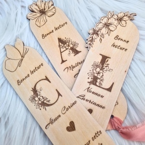 May include: Wooden bookmarks with floral designs and engraved letters. The bookmarks have the words "Bonne lecture" and names engraved on them. The bookmarks are light brown and have a natural wood grain.