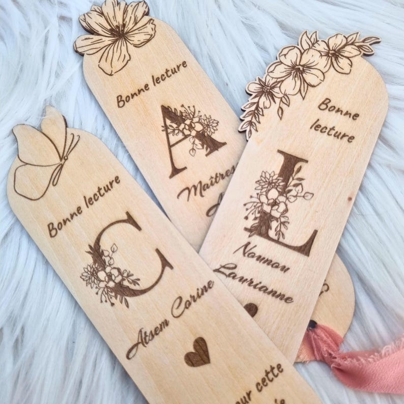 May include: Wooden bookmarks with floral designs and engraved letters. The bookmarks have the words "Bonne lecture" and names engraved on them. The bookmarks are light brown and have a natural wood grain.