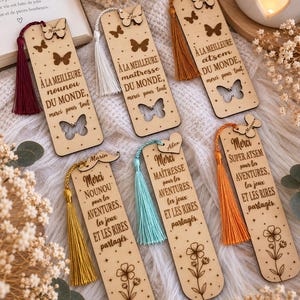 Peut inclure: Six marque-pages rectangulaires en bois aux coins arrondis, gravés au laser avec des textes en français, des motifs de papillons ou de fleurs, et des pompons colorés. Les prénoms Nina, Lea, Naty, Marin, Alex et Manon sont inscrits avec des découpes.