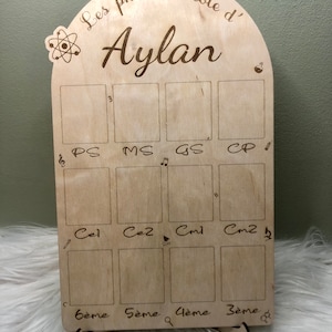 Peut inclure: Un présentoir photo en bois avec une forme d'arche découpée et le texte "Les photos d'école d' Aylan". Le présentoir comporte 10 espaces rectangulaires pour photos, étiquetés avec les classes d'école françaises : PS, MS, GS, CP, Ce1, Ce2, Cm1, Cm2, 6ème, 5ème, 4ème et 3ème.