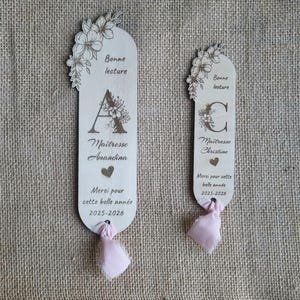 May include: Two wooden bookmarks with floral designs and text. One bookmark features the letter "A" and the name "Amandina", the other has the letter "C" and the name "Christine". Both have pink ribbons.