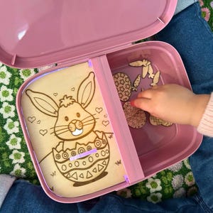 Peut inclure: Une boîte de rangement en plastique rose avec un panneau en bois représentant un lapin de dessin animé à l'intérieur d'un œuf. Le panneau est éclairé par une lumière violette. La boîte contient des découpes en bois d'œufs de Pâques et de carottes.