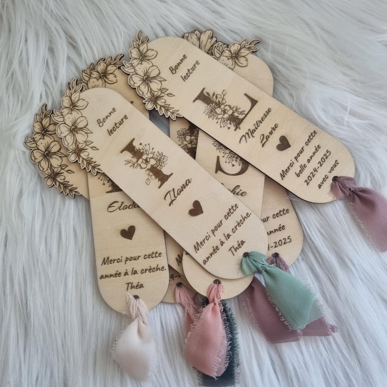 May include: Wooden bookmarks with floral designs and personalized text. Each bookmark features a decorative floral border, an initial, and a ribbon. The text includes names, phrases like "Bonne lecture," and dates.