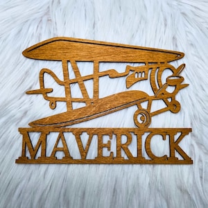 Peut inclure: Une pancarte en bois avec une découpe d'un biplan et le nom "Maverick" en dessous. La pancarte est teintée d'une couleur brun clair.