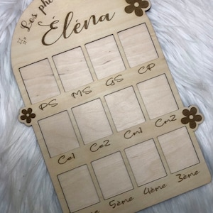 Peut inclure: Un cadre photo en bois avec une forme d'arche découpée et un motif floral. Le cadre a des espaces pour des photos avec des étiquettes pour chaque niveau scolaire de PS à 3ème. Le cadre est intitulé "Les photos d'école d'Elena".