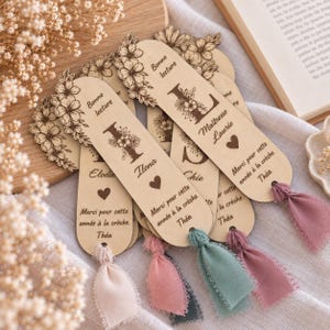 May include: A collection of light-colored wooden bookmarks with rounded ends, each featuring engraved floral designs, a large initial, and the text "Bonne lecture" and "Merci pour cette ann&eacute;e &agrave; la cr&egrave;che." Each bookmark includes a frayed fabric tassel.