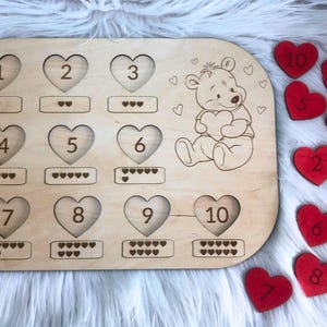 Può includere: Giocattolo di apprendimento in legno con un'illustrazione di un orso e pezzi a forma di cuore numerati da 1 a 10. Il giocattolo include un gioco di abbinamento con file di cuori per ogni numero.