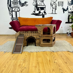 K&ouml;nnte beinhalten: Ein h&ouml;lzernes Katzenspielhaus mit einer Rampe, einem Tunnel und zwei Ebenen. Das Spielhaus ist braun und hat eine nat&uuml;rliche Holzoptik. Es steht auf einem hellbraunen Teppich vor einem roten Sofa.