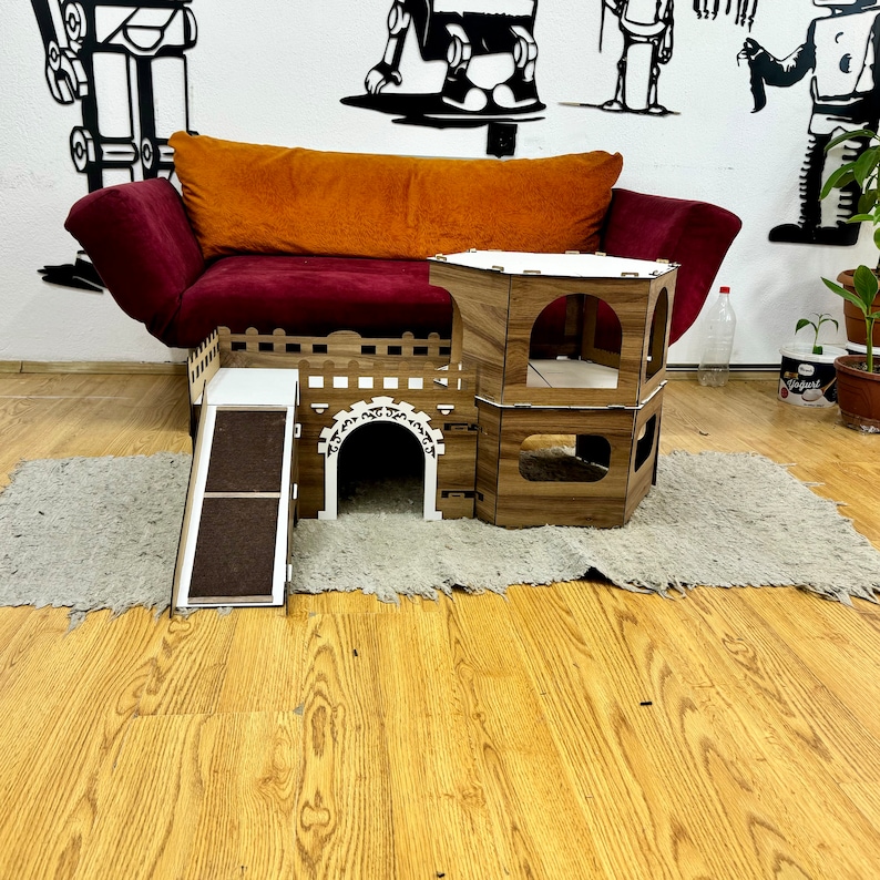 K&ouml;nnte beinhalten: Ein Katzenhaus aus Holz mit einer Rampe, einem Tunnel und einem Turm. Das Haus ist in hellbraun lackiert und hat ein wei&szlig;es Dach. Das Haus steht auf einem wei&szlig;en Teppich vor einer braunen Couch.