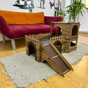 K&ouml;nnte beinhalten: Ein h&ouml;lzernes Katzenspielhaus mit einer Rutsche, einem Tunnel und mehreren Ebenen. Das Spielhaus ist braun und hat eine nat&uuml;rliche Holzoptik. Es steht auf einem hellbraunen Teppich vor einem roten Sofa.