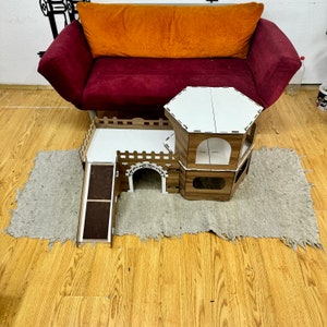 K&ouml;nnte beinhalten: Ein mehrst&ouml;ckiges Haus aus Holz f&uuml;r Haustiere mit einer Rampe, einem wei&szlig;en Dach und einer braunen und wei&szlig;en Farbgebung. Das Haus steht auf einem grauen Teppich vor einem roten Sofa.