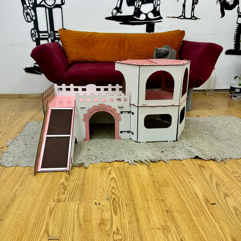 K&ouml;nnte beinhalten: Ein rosa-wei&szlig;es Katzenspielhaus mit einer Rampe und einem Turm. Das Spielhaus ist aus Holz und hat einen Ausschnitt in Form einer Burgt&uuml;r. Das Spielhaus steht auf einem hellbraunen Holzboden.