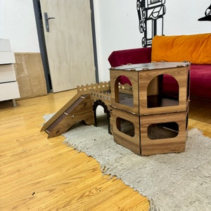 K&ouml;nnte beinhalten: Ein h&ouml;lzernes Katzenspielhaus mit einer Rampe und zwei Ebenen. Das Spielhaus ist braun und hat eine sechseckige Form. Das Spielhaus steht auf einem hellbraunen Teppich.