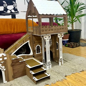 K&ouml;nnte beinhalten: Ein h&ouml;lzernes Katzenspielhaus mit einer Rampe, einem Aussichtsturm und einer dreistufigen Leiter. Das Spielhaus ist braun und wei&szlig; gestrichen und hat ein wei&szlig;es Dach. Eine Katze schaut aus einem Loch in der Seite des Spielhauses.