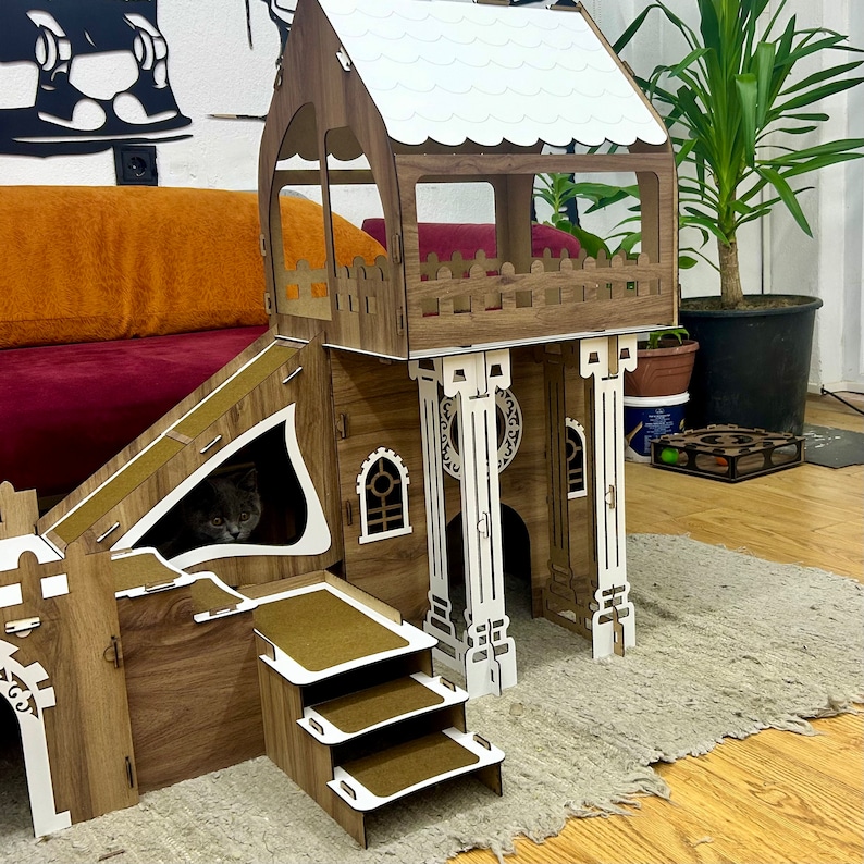 K&ouml;nnte beinhalten: Ein h&ouml;lzernes Katzenspielhaus mit einer Rampe, einem Aussichtsturm und einer dreistufigen Leiter. Das Spielhaus ist braun und wei&szlig; gestrichen und hat ein wei&szlig;es Dach. Eine Katze schaut aus einem Loch in der Seite des Spielhauses.