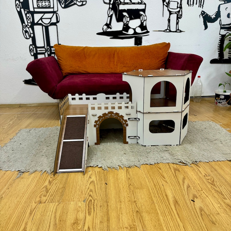 K&ouml;nnte beinhalten: Ein wei&szlig;es und braunes Holzkatzenhaus mit einer Rampe und einem Turm. Das Haus hat einen kleinen Eingang und eine gro&szlig;e &Ouml;ffnung oben. Das Haus steht auf einem hellbraunen Teppich.