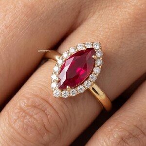 May include: A gold ring featuring a marquise-cut, deep red gemstone surrounded by a halo of smaller, clear stones. The ring's band is a simple, polished gold. The ring is shown on a person's finger.