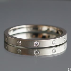 Op de afbeelding: Twee zilveren ringen met kleine, gelijkmatig verdeelde, heldere edelstenen. De ringen worden getoond op een reflecterend oppervlak, met de merknaam "gemstreets".