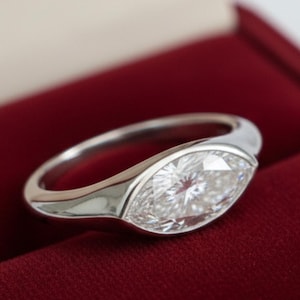 Può includere: Anello con diamante taglio marquise e fascia in oro bianco, presentato in un cofanetto di velluto rosso. Il diamante, punto focale, riflette la luce. Un gioiello elegante per un'occasione speciale.