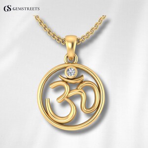 May include: A gold-colored pendant necklace featuring the Om symbol. The pendant has a circular design with the Om symbol in the center, and a small, clear gemstone is set at the top. The necklace chain is also gold-colored.