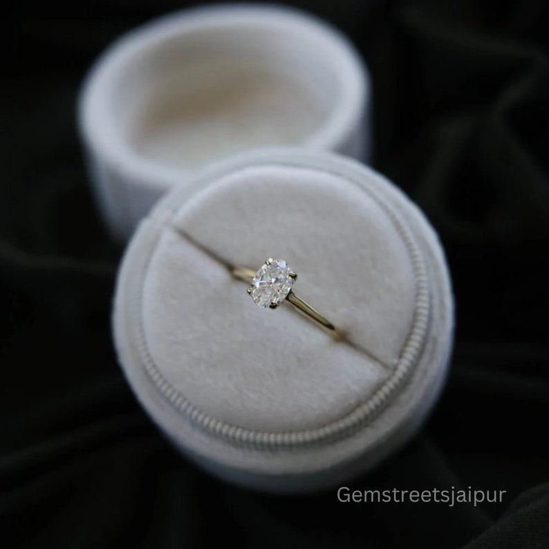 May include: A gold ring with an oval-shaped diamond in a white ring box. The ring is set in a simple band.