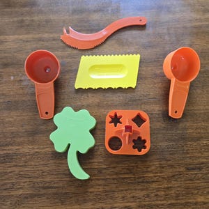 Peut inclure: Ensemble d'ustensiles de cuisine colorés, comprenant des tasses à mesurer oranges, une raclette jaune et des emporte-pièces verts et oranges. Les outils sont disposés sur une surface en bois, mettant en valeur leurs formes et fonctions.