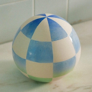 Può includere: Oggetto decorativo sferico con un motivo a scacchi in azzurro e bianco. Il bordo inferiore ha una fascia verde. L'oggetto è probabilmente in ceramica o porcellana e poggia su una superficie bianca.
