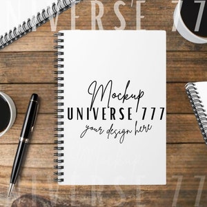 May include: A white spiral bound notebook with the text "Mockup UNIVERSE 777 your design here" on the front cover. The notebook is on a wooden table with a black pen and two cups of coffee.